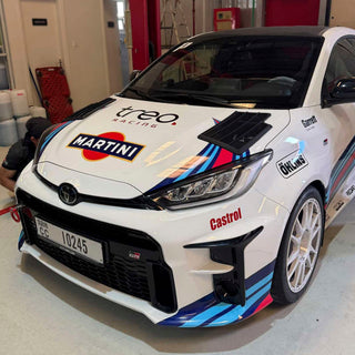 White GR Yaris race car with various sponsor logos in a garage setting, with PIRAS Motorsport Rally.1 Hood vents